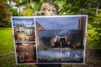 plansza z wizualizacją projektu architektonicznego w kolorach szaroniebieskim i rudym, trzymana przez młodą dziewczyną stojącą w parku